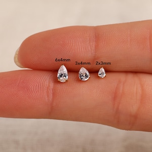 May include: Three pear-shaped clear stud earrings in gold settings, arranged by size. The labels indicate dimensions of 6x4mm, 3x4mm, and 2x3mm. These small, faceted stones are displayed in a row, showcasing their teardrop shape and clear, reflective appearance.