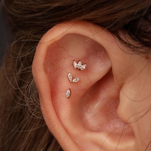 May include: Three gold-toned flat back earrings arranged vertically along the ear cartilage. The top earring features three marquise-cut clear stones, the middle has two, and the bottom earring consists of a single marquise-cut clear stone.