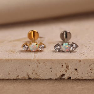 May include: Two flat-back labret studs featuring a central round opal flanked by two smaller clear round stones. One stud has a gold-tone metal finish, while the other has a silver-tone metal finish. Both pieces feature a circular base post.