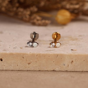 May include: Two labret studs featuring a flat back, a straight post, and a decorative front with one clear round gemstone and one small white round stone. One stud is silver-toned metal, and the other is gold-toned metal, both displayed upright.