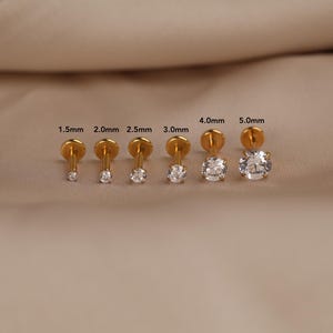 May include: A set of six gold-tone flat back stud earrings with clear round-cut stones, arranged in increasing sizes from 1.5mm to 5.0mm. Each stud features a post and a circular flat backing, with the specific size labeled above each piece.