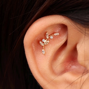 May include: A gold-toned ear cartilage piercing jewelry piece shaped like a curved branch with multiple marquise-cut and round-cut clear white stones arranged along the stem, secured within the upper ear cartilage area.