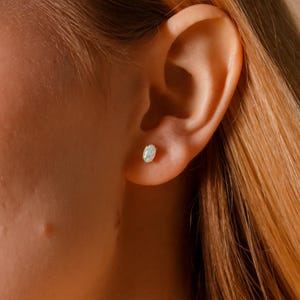 May include: A close-up of an ear with a small, oval-shaped stud earring. The earring has a light-colored, possibly iridescent stone set in a gold-colored metal. The skin around the ear is fair-toned, and the hair is a light shade of blonde.