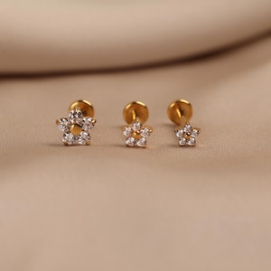 May include: Three gold-tone flat back stud earrings in varying sizes, each shaped like a flower with five petals. Each petal is set with a small clear stone, and a small gold-tone bead is positioned in the center of each flower-shaped earring head.