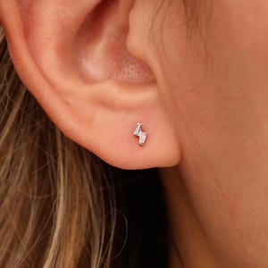May include: A small, silver-toned stud earring shaped like a lightning bolt. The earring features two clear, baguette-cut stones set within a metallic frame, positioned securely in the earlobe. The design is simple, geometric, and reflective.