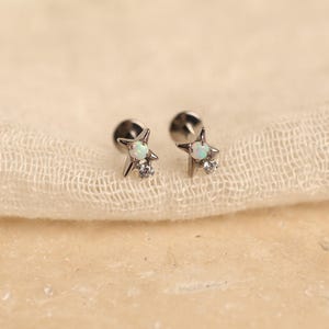 May include: A pair of silver-toned star-shaped earrings. Each earring features a small, white opal stone at the center of the star and a clear, round gemstone below. The earrings are displayed on a cream-colored, textured fabric.