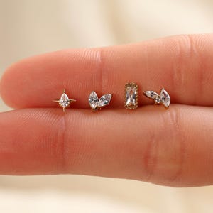 May include: Four gold-tone stud earrings featuring clear, faceted stones. The designs include a star-shaped setting with a pear-cut stone, a double marquise-cut stone cluster, a rectangular baguette-cut stone with a beaded border, and a small wing-like cluster.