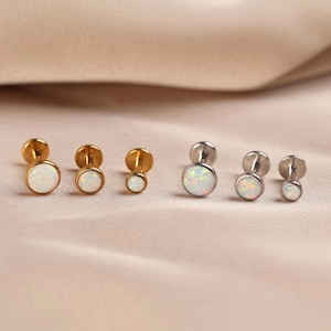 May include: A collection of six flat-back stud earrings arranged in two rows of three. Three studs feature gold-tone metal settings, and three feature silver-tone metal settings. Each stud is topped with a round, white, iridescent opal-like stone.