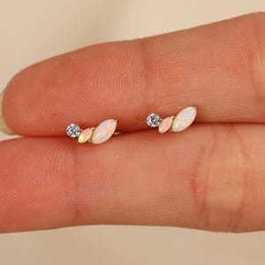 May include: A pair of gold stud earrings, each featuring a small round clear stone, a small marquise-shaped opal, and a larger marquise-shaped opal arranged in a curved, cluster design. The earrings are crafted with a yellow gold metal setting.