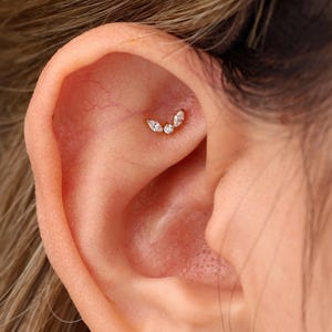 May include: A gold-toned ear piercing stud featuring a curved design with two marquise-cut clear stones flanking a single round-cut clear stone in the center. The jewelry is positioned in the upper cartilage area of the ear, showcasing a delicate metal setting.