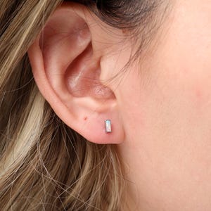 May include: A close-up of an ear with a small, rectangular earring. The earring has a silver-colored setting and a light-colored, iridescent stone. The person's hair is blonde with highlights.