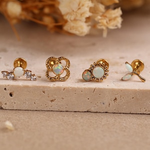 May include: Four gold-tone flat back stud earrings featuring opal and clear crystal accents. The designs include a curved bar with crystals, a four-leaf clover shape, a circular sunburst pattern with two opals, and a small three-leaf plant motif.