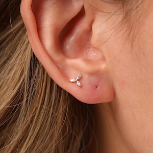 May include: A small gold stud earring featuring three marquise-cut clear stones arranged in a delicate floral or leaf-like cluster. The earring is positioned in the lower earlobe, showcasing a metallic gold setting and sparkling white stones.
