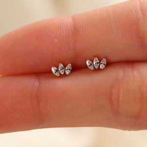 May include: A pair of silver-toned stud earrings, each featuring three small, clear marquise-cut stones arranged in a fan-like cluster. The earrings are metallic with a polished finish, showcasing a delicate and symmetrical design with pointed edges.