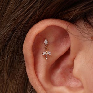 May include: A gold-toned cartilage earring featuring a single marquise-cut clear stone at the top, connected by a small jump ring to a dangling cluster of three marquise-cut clear stones arranged in a fan-like shape, secured in the ear.