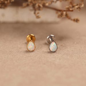 May include: Two labret studs featuring teardrop-shaped white opal stones. One stud has a gold-toned metal setting and post, while the other has a silver-toned metal setting and post. Both pieces display iridescent flashes of color within the stones.