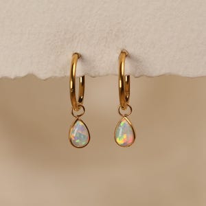 May include: A pair of gold hoop earrings featuring dangling teardrop-shaped opal charms. Each earring consists of a polished gold hoop with a small jump ring connecting to a bezel-set, iridescent white opal stone that displays flashes of color.