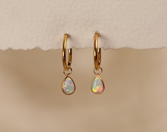 May include: A pair of gold hoop earrings featuring dangling teardrop-shaped opal charms. Each earring consists of a polished gold hoop with a small jump ring connecting to a bezel-set, iridescent white opal stone that displays flashes of color.
