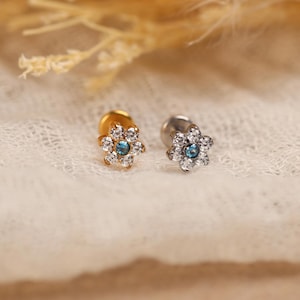 May include: Two flower-shaped stud earrings featuring a central blue stone surrounded by six clear white stones. One earring has a gold-tone metal base, while the other has a silver-tone metal base, both with flat circular backings.