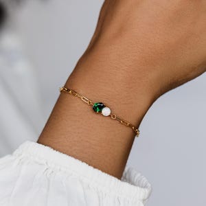 Könnte beinhalten: Ein goldfarbenes Kettenarmband mit einem kleinen Cluster aus grünen und weißen Edelsteinen. Das Armband wird am Handgelenk getragen, der Hintergrund ist ein weiches Weiß. Ein zarter Schmuck.