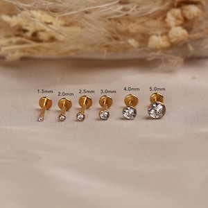 May include: A set of six gold-tone flat back labret studs with clear round gemstones, arranged in increasing size from 1.5mm to 5.0mm. Each stud features a circular metal base and a prong-set stone, with the specific size labeled above each piece.