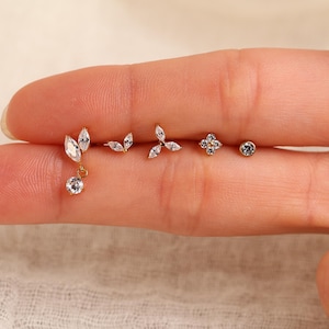 May include: A set of five gold-tone stud earrings featuring clear, faceted cubic zirconia stones. The designs include a marquise-cut drop, a two-leaf marquise cluster, a three-leaf marquise cluster, a four-petal flower, and a single round stone.
