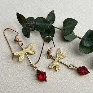 Puede incluir: Pendientes dorados con forma de libélula y gemas rojas en forma de diamante. Los pendientes presentan colgantes detallados de libélula y están suspendidos de delicados ganchos para las orejas. Una ramita de hojas verdes proporciona un fondo natural.