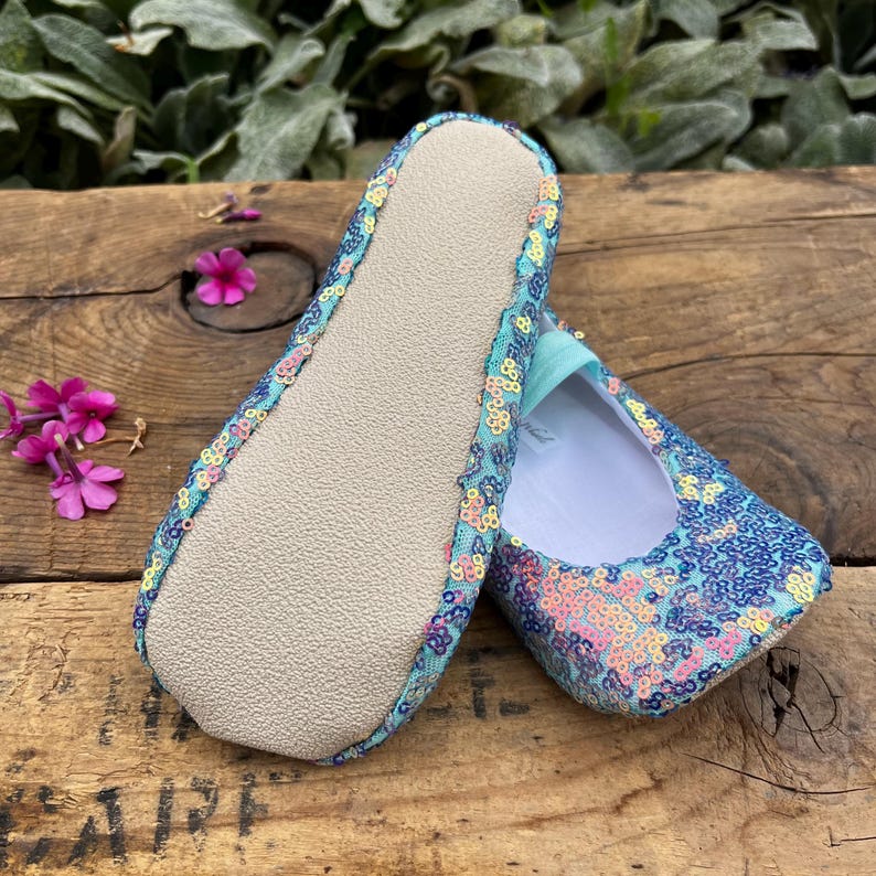 May include: A pair of slip-on shoes featuring a light blue fabric base covered in iridescent, multi-colored sequins. One shoe is positioned to show the textured, light-colored sole, while the other displays the interior with a light blue elastic strap.