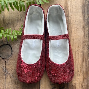 May include: A pair of red sequined Mary Jane style slippers with white lining. The slippers have a strap across the top and are displayed on a rustic wooden surface. The slippers are a festive, eye-catching footwear.