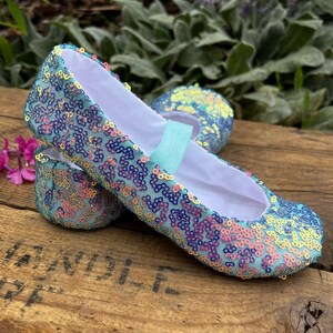 May include: A pair of ballet flats covered in iridescent sequins that shift between shades of blue, purple, and pink. The shoes feature a white interior lining and a light blue elastic strap across the top of the foot opening for a secure fit.