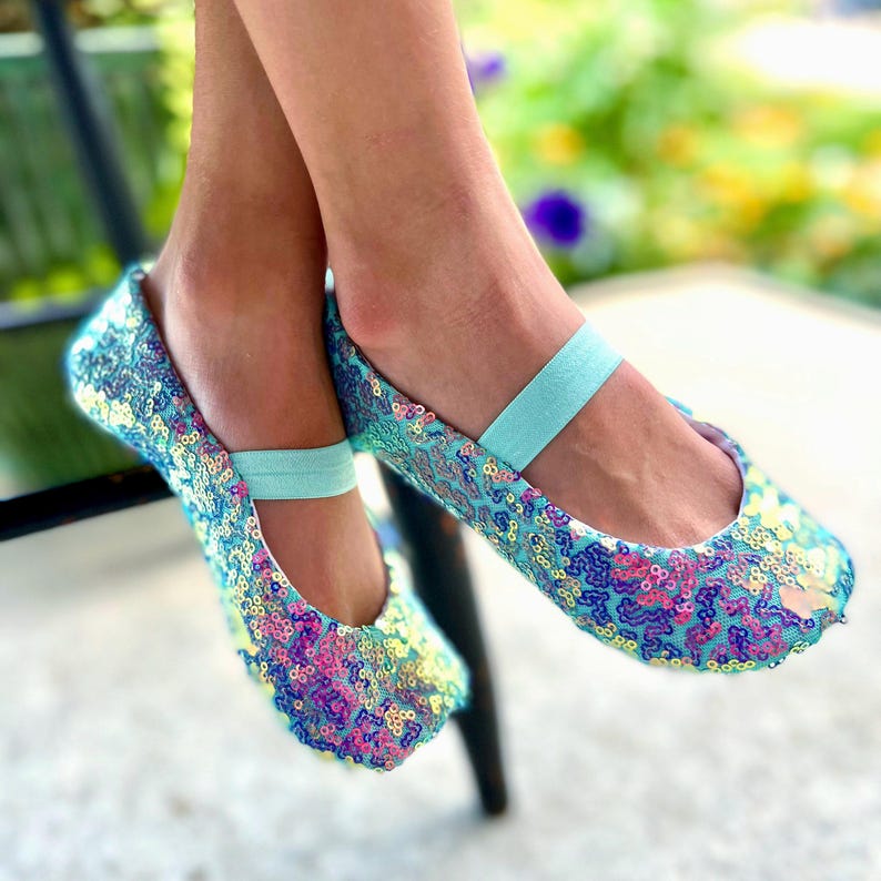 May include: A pair of ballet flats covered in iridescent sequins in shades of blue, pink, and yellow. The shoes feature a light blue elastic strap across the top of the foot, providing a secure fit. The sequined material creates a shimmering texture.