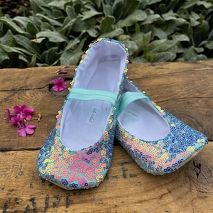 May include: A pair of flat shoes covered in iridescent sequins that shift between shades of blue, pink, and yellow. Each shoe features a light blue elastic strap across the top and a white interior lining with a small white label reading Titz Whitz.