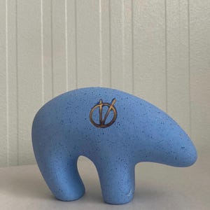 May include: A light blue bear figurine with a speckled texture and a dark brown circular symbol featuring a vertical line and a V-shape etched into the side. The sculpture has a smooth, rounded form and stands on two thick, sturdy legs.