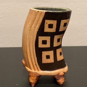 May include: A handcrafted ceramic vase with a unique, curved shape. The vase features a tan exterior with a black panel adorned with tan square designs. It rests on three small, brown feet and has a green interior.