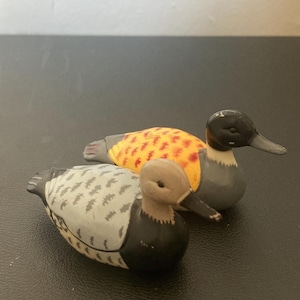 May include: Two small duck figurines. The front duck has a light gray head, black chest, and a light gray back with dark gray feather patterns. The rear duck has a black head, gray chest, and a yellow back with orange feather markings.
