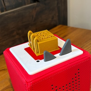 May include: A red cube-shaped speaker with a white top and a black speaker grille. The speaker has two gray cat ears on top. A small gold-colored object with the text "BOOK OF MORMON" is on top of the speaker.