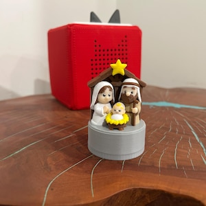 May include: A small, detailed nativity scene figurine featuring Mary, Joseph, and baby Jesus in a manger, topped with a yellow star. The figurine sits on a gray base, with a red cube-shaped object in the background.