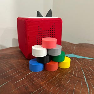 May include: A red fabric-covered audio player with black ears and a speaker pattern. A stack of colorful, round, plastic lids in various colors, including red, white, black, gray, green, blue, orange, and yellow, are in front of the player.