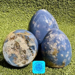May include: Three blue decorative stone eggs of varying sizes. The eggs have a mottled appearance with lighter blue and brown patches. The eggs are arranged on a green surface. A small, square logo with the text "Glittering Grit" is in the bottom right corner.