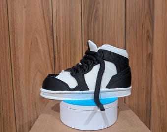 May include: A small, black and white high-top sneaker with black laces. The shoe has a white sole and is displayed on a white rotating platform with a blue light. The background is a wooden wall.