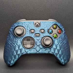 May include: A blue and black Xbox controller with a textured, scaly design. The controller has a standard layout with buttons, joysticks, and a directional pad.