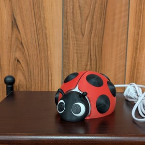 May include: A red and black ladybug-shaped night light with a white power cord and switch. The ladybug has large, white-rimmed eyes and black spots. It sits on a dark brown table against a wood-paneled wall.