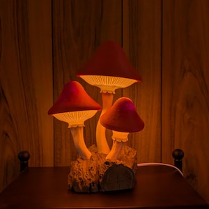 May include: A whimsical mushroom lamp with three glowing red caps and white stems. The lamp is set on a wooden base and emits a warm, inviting light. The background is a wooden wall.
