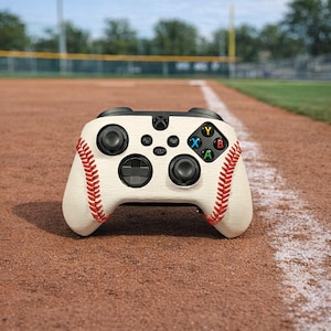 May include: A baseball-themed Xbox controller with a cream-colored body and red stitching, resembling a baseball. The controller features black buttons and directional pads, with colored letter buttons. The controller is resting on a baseball field.