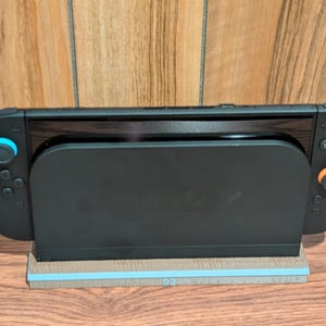 May include: A black Nintendo Switch console docked in a black charging station. The console has blue and orange controllers. The charging station sits on a wooden base with a blue stripe.