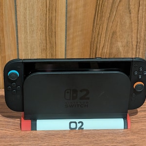 May include: Black Nintendo Switch console docked in a black stand with the text "02 Nintendo Switch". The stand has a red and light blue base. The console has blue and orange controllers.