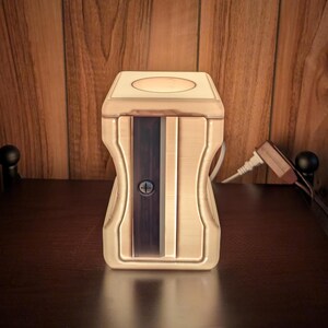 May include: A large, light-colored, rectangular pencil sharpener with a central dark metal blade assembly secured by a single visible screw. The device features a circular opening on the top surface and a contoured, hourglass-shaped body design.