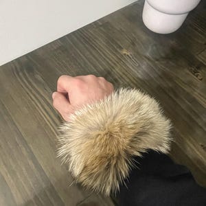 May include: A close-up of a black sleeve with a light brown fur cuff. The fur is thick and fluffy, with a mix of light and dark brown tones. The sleeve is made of a dark material, possibly wool or a similar fabric.