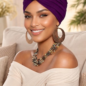 May include: A woman wearing a deep purple turban with a knotted top. She is also wearing large, circular, brown earrings and a multi-strand beaded necklace. Her off-the-shoulder top is a light beige color.