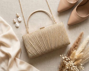 May include: A gold clutch with a chain strap and a matching handle, paired with beige heels and pearl earrings. The clutch has a textured, pleated design. A bouquet of dried flowers and a piece of silk fabric complete the elegant arrangement.